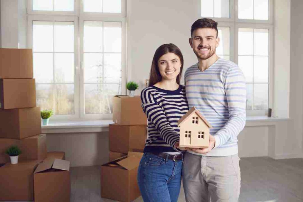 Smiling couple holding a wooden house model in their new home surrounded by boxes.
