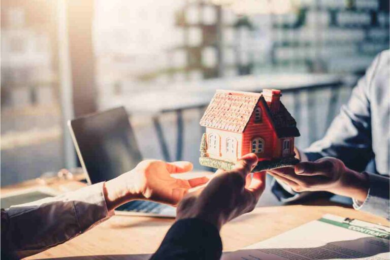 Hands exchanging a red-roofed model house over a desk with documents.